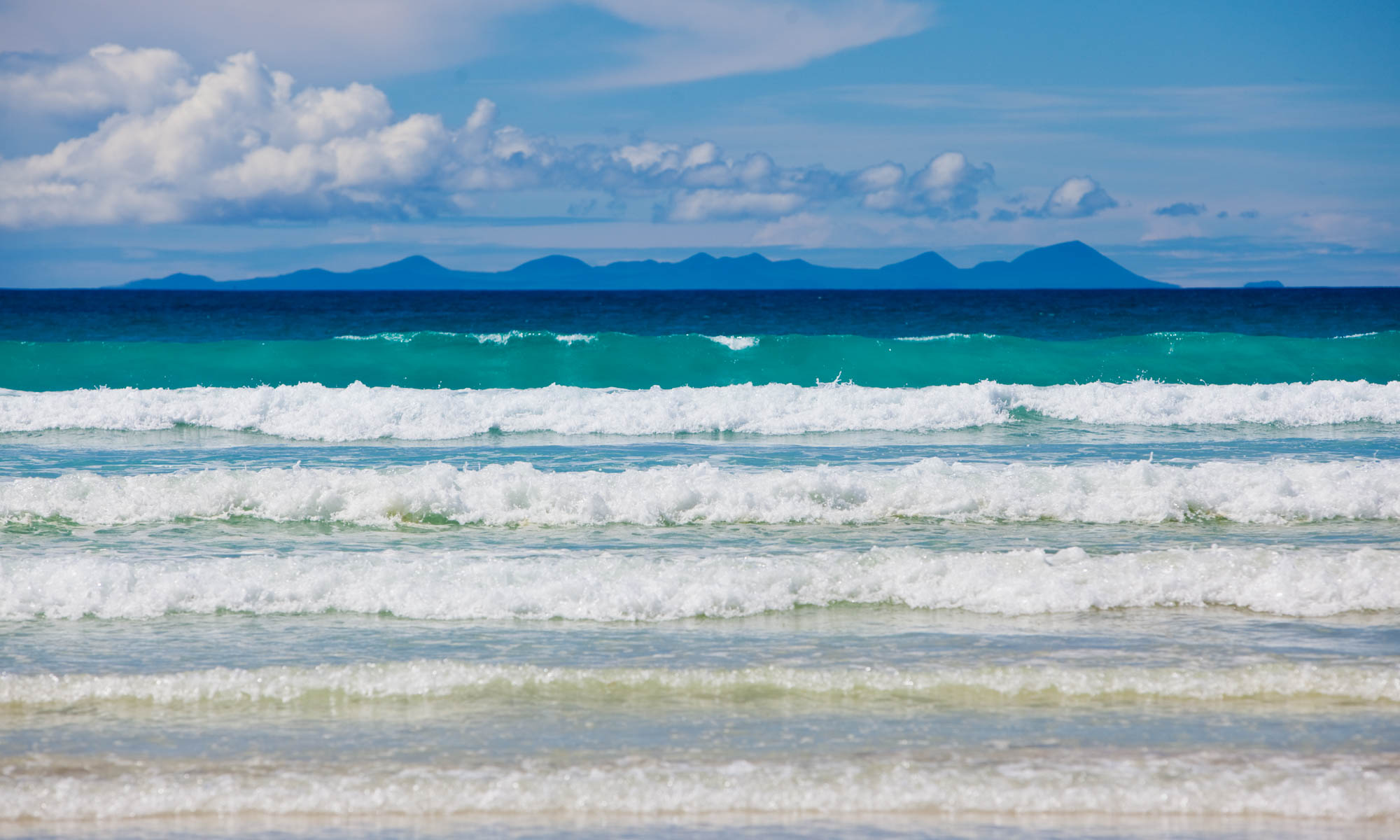 Beautiful waves in various shades of blue rolling into a beach in the Galapagos Islands with Floreana Island in the distance.