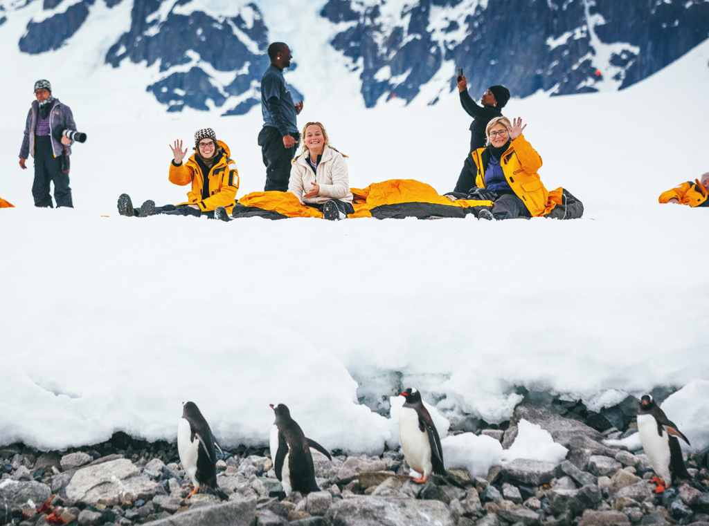 three women sitting on the snow and smiling at the camera with penguins in the foreground